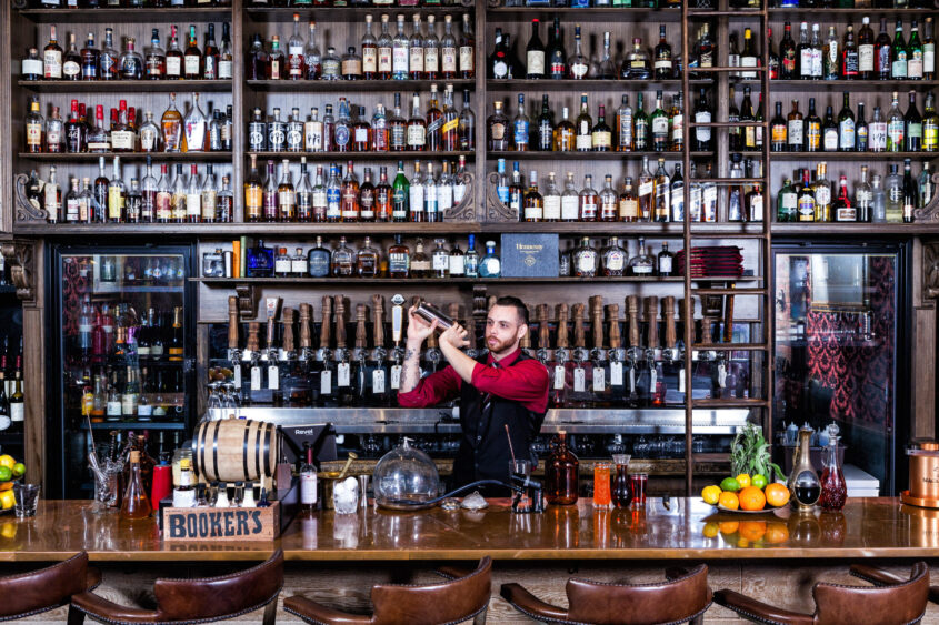A bartender in a red shirt and black apron expertly shakes a cocktail shaker, his wrist frequency on display behind a bar lined with bottles. The shelves behind him are filled with liquor bottles, glasses, and bar tools. Fruits and bottles sit on the counter. Lifestyle