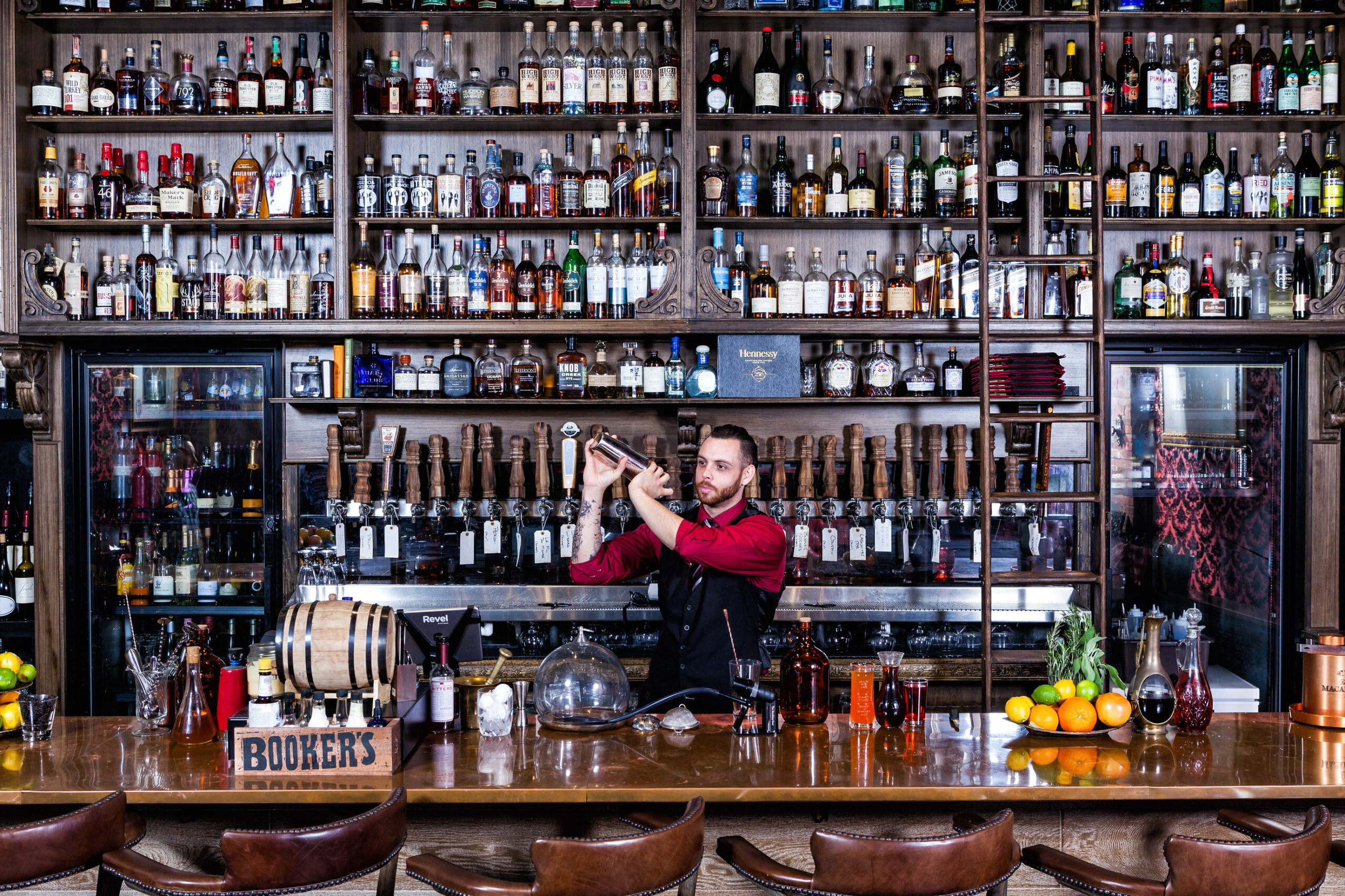 A bartender in a red shirt and black apron expertly shakes a cocktail shaker, his wrist frequency on display behind a bar lined with bottles. The shelves behind him are filled with liquor bottles, glasses, and bar tools. Fruits and bottles sit on the counter. Lifestyle