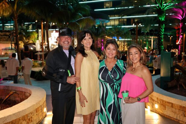 Four adults pose and smile together at an outdoor evening event with palm trees, colorful lights, and a lively crowd in the background. Dressed in semi-formal attire and holding green cups, they radiate Summer Vibes. Lifestyle