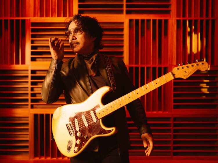 A man wearing glasses, a scarf, and a leather jacket holds an electric guitar in front of geometric wooden panels, bathed in warm orange lighting—a scene reminiscent of classic Hall & Oates album covers. Lifestyle