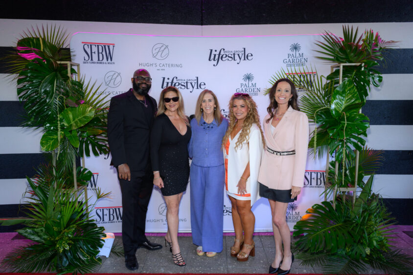 Five people stand smiling in front of a backdrop with logos and tropical plants at the Palm Garden Event House, dressed in formal and semi-formal attire for the March Cover Party social or networking event. Lifestyle