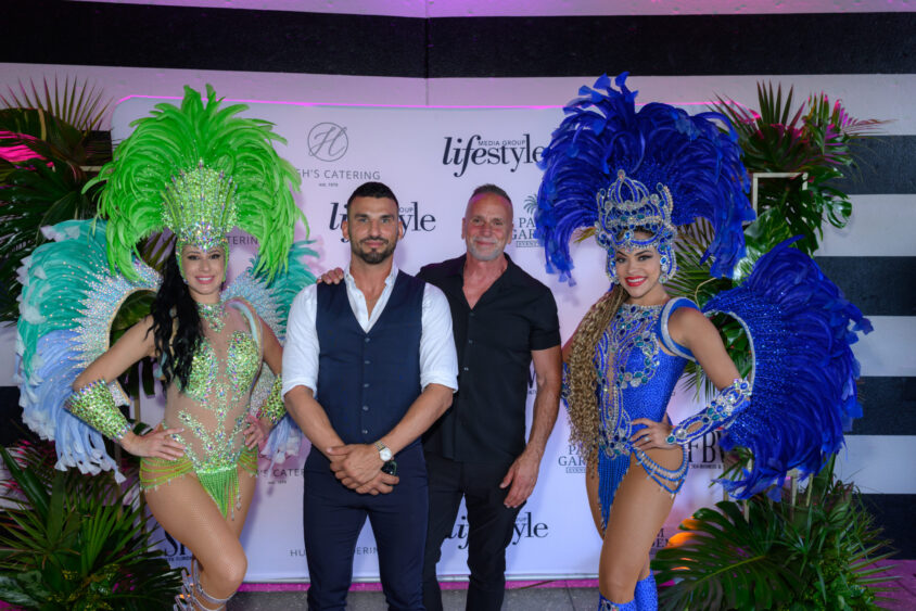 Two men pose between two women in elaborate feathered costumes, one in green and the other in blue, at the March Cover Party hosted by Palm Garden Event House, with palm leaves framing the branded event backdrop. Lifestyle