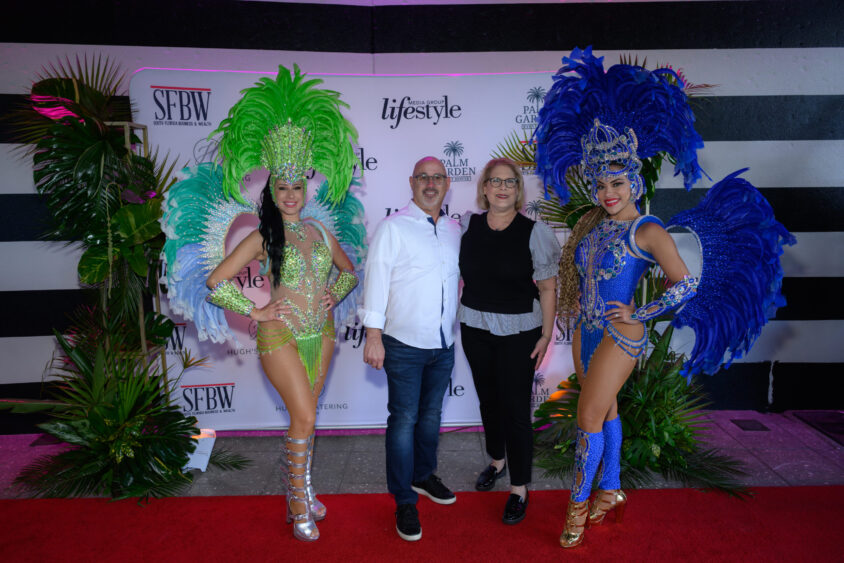 Two performers in bright feathered costumes pose with a man and a woman on a red carpet at the March Cover Party, held at Palm Garden Event House, with palm plants and a "Lifestyle" backdrop behind them. Lifestyle