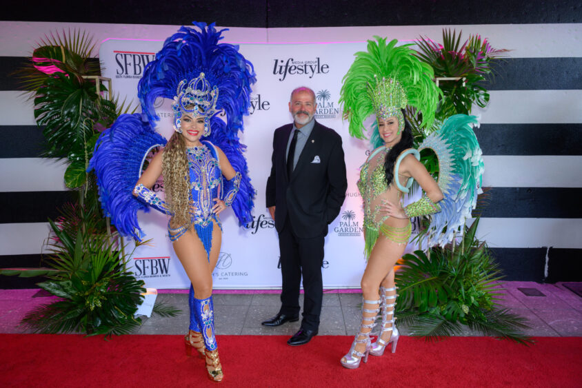 A man in a suit stands on a red carpet at the March Cover Party between two women in elaborate blue and green feathered showgirl costumes, set against a backdrop with logos and tropical plants at Palm Garden Event House. Lifestyle