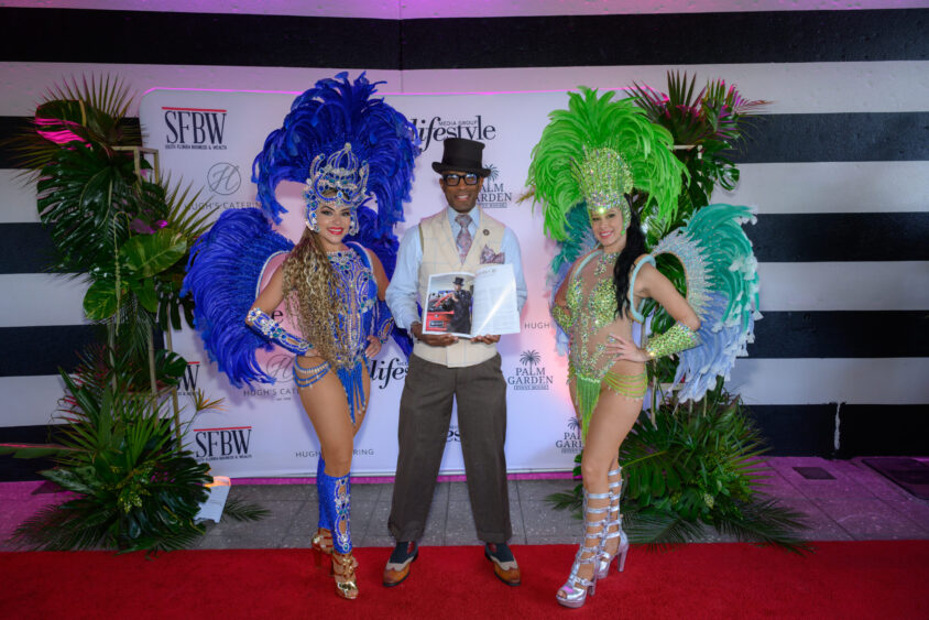 Three people pose on the red carpet at the Palm Garden Event House March Cover Party: a man in a suit, sunglasses, and hat holds a magazine between two women in vibrant blue and green feathered showgirl costumes, framed by event logos and tropical plants. Lifestyle