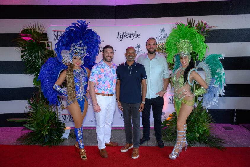 Four men stand on a red carpet between two women in showgirl costumes with feathered headdresses, one in blue and one in green, at the Palm Garden Event House March Cover Party, posing before a step-and-repeat backdrop with tropical plants. Lifestyle