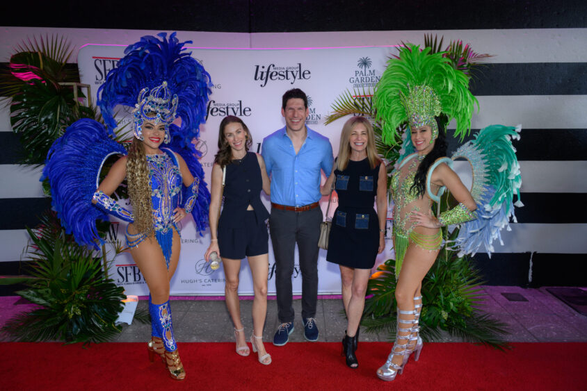 Four people pose on a red carpet at the Palm Garden Event House March Cover Party—two women in feathered showgirl costumes stand on each end, while a man and two women in casual attire smile in front of a branded backdrop. Lifestyle