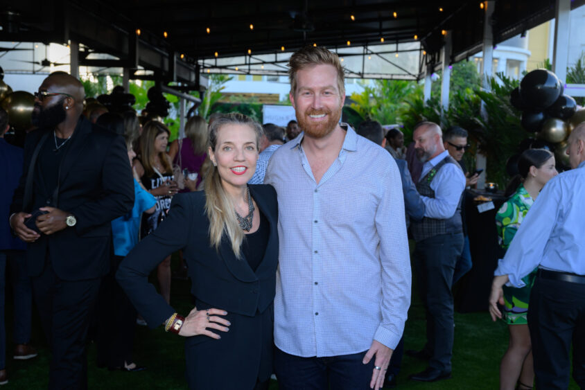 A smiling woman in a black outfit stands next to a smiling man in a light shirt at the March Cover Party, with guests mingling in the background under the covered area at Palm Garden Event House. Lifestyle