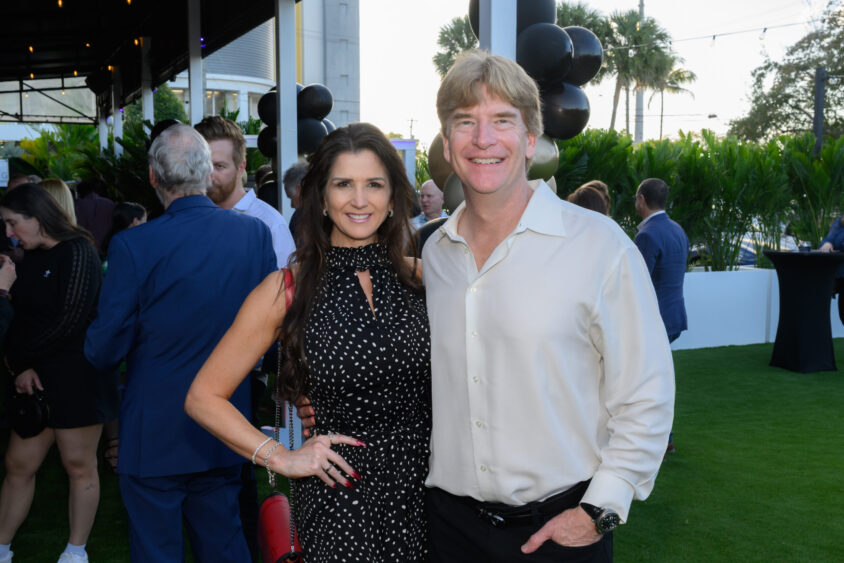 A smiling couple poses together at the March Cover Party held at Palm Garden Event House, with green grass, palm trees, and black balloons in the background. The woman wears a black dress with white polka dots, and the man is in a light-colored shirt. Lifestyle