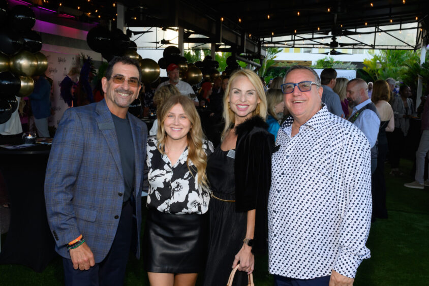 Four people stand together smiling at an outdoor March Cover Party with string lights, greenery, and balloons at Palm Garden Event House. The group is dressed in semi-formal attire and appears to be enjoying a lively social gathering. Lifestyle