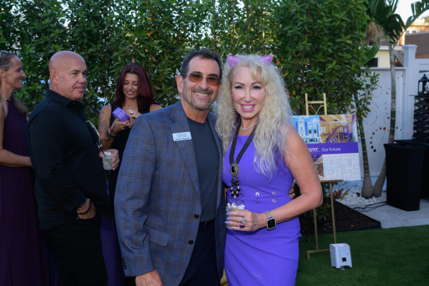 A man in a blue plaid jacket and a woman with long blonde hair, cat ears, and a black dress smile at the Palm Garden Event House’s March Cover Party, with other guests and a poster in the background. Lifestyle