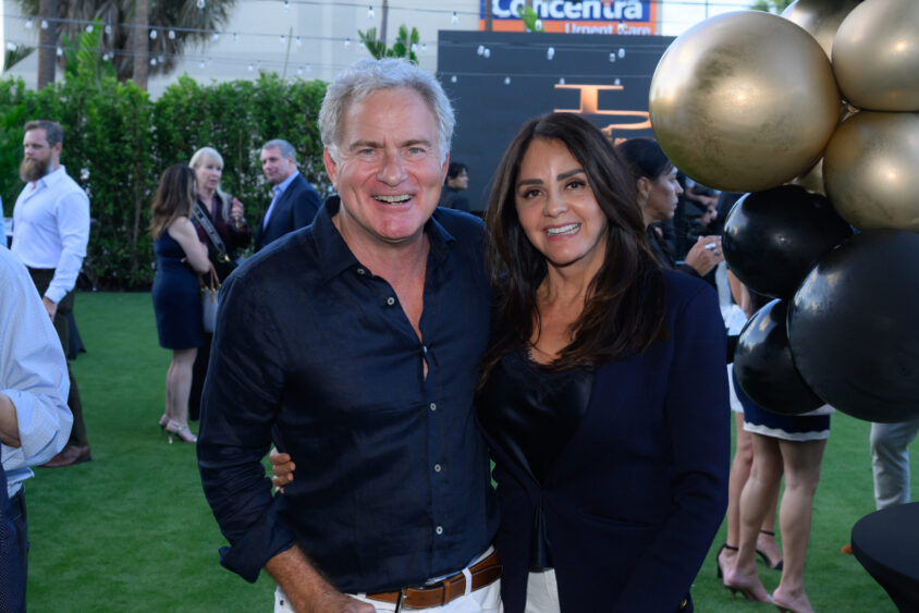 A smiling man and woman pose together at the March Cover Party, held outdoors at Palm Garden Event House. They stand next to black and gold balloons on a grassy area with trees and a building in the background. Lifestyle