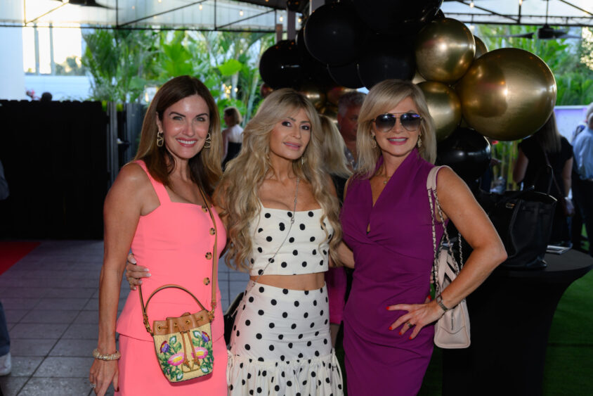 Three women pose together at the March Cover Party at Palm Garden Event House. One wears a pink dress with a floral handbag, the second sports a white polka dot outfit, and the third dons a purple dress and sunglasses amid gold and black balloons. Lifestyle