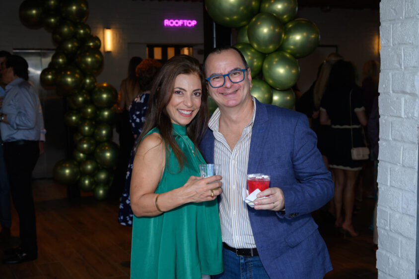 A woman in a green dress and a man in a blue jacket smile and pose with drinks at the March Cover Party held at Palm Garden Event House, where green balloons decorate the venue and other guests mingle in the background. Lifestyle