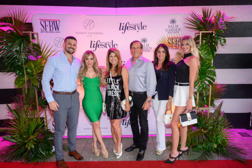 Six people pose and smile on a red carpet at the Palm Garden Event House’s March Cover Party, in front of a step-and-repeat backdrop with magazine and sponsor logos, surrounded by green plants and lit by pink lighting. Lifestyle
