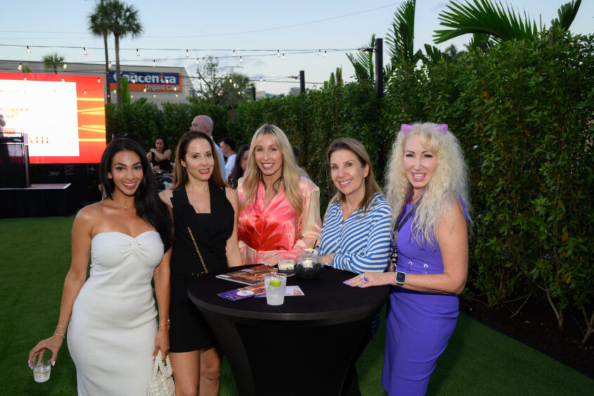 Five women pose and smile around a tall cocktail table outdoors at the Palm Garden Event House during the March Cover Party. Palm trees, string lights, and greenery surround them as they enjoy drinks in colorful dresses by a large red sign. Lifestyle