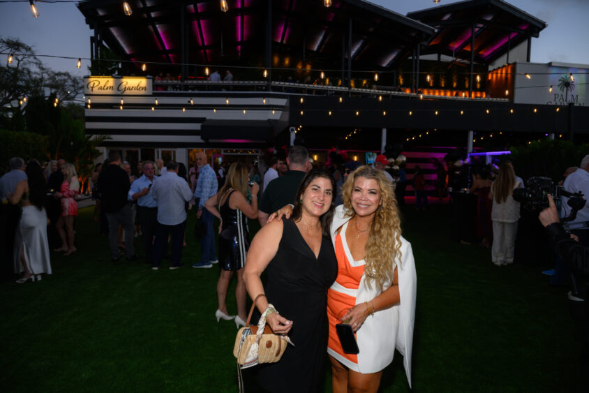 Two women pose and smile at the March Cover Party, an outdoor evening event at Palm Garden Event House, with a crowd and string lights in the background. The woman on the left wears a black dress; the woman on the right wears orange and white with a jacket. Lifestyle