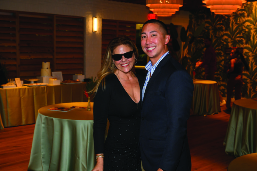 A woman in a black dress and sunglasses stands next to a smiling man in a suit at the March Cover Party, posing indoors at Palm Garden Event House with round tables and soft lighting in the background. Lifestyle