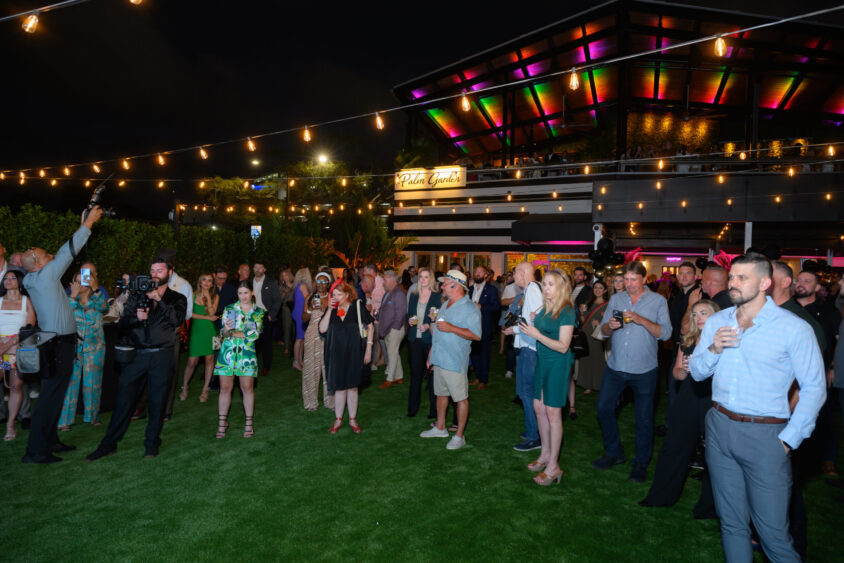 A large group at the Palm Garden Event House’s March Cover Party stand on grass, watching something off camera. String lights hang overhead, and a modern building with colorful lights is in the background. Most guests are dressed in semi-formal attire. Lifestyle