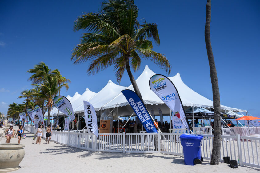 White event tents with sponsor flags line Hollywood Beach under clear blue skies and palm trees, as people stroll by and a blue recycling bin sits in the foreground at SAVOR SoFlo. Lifestyle
