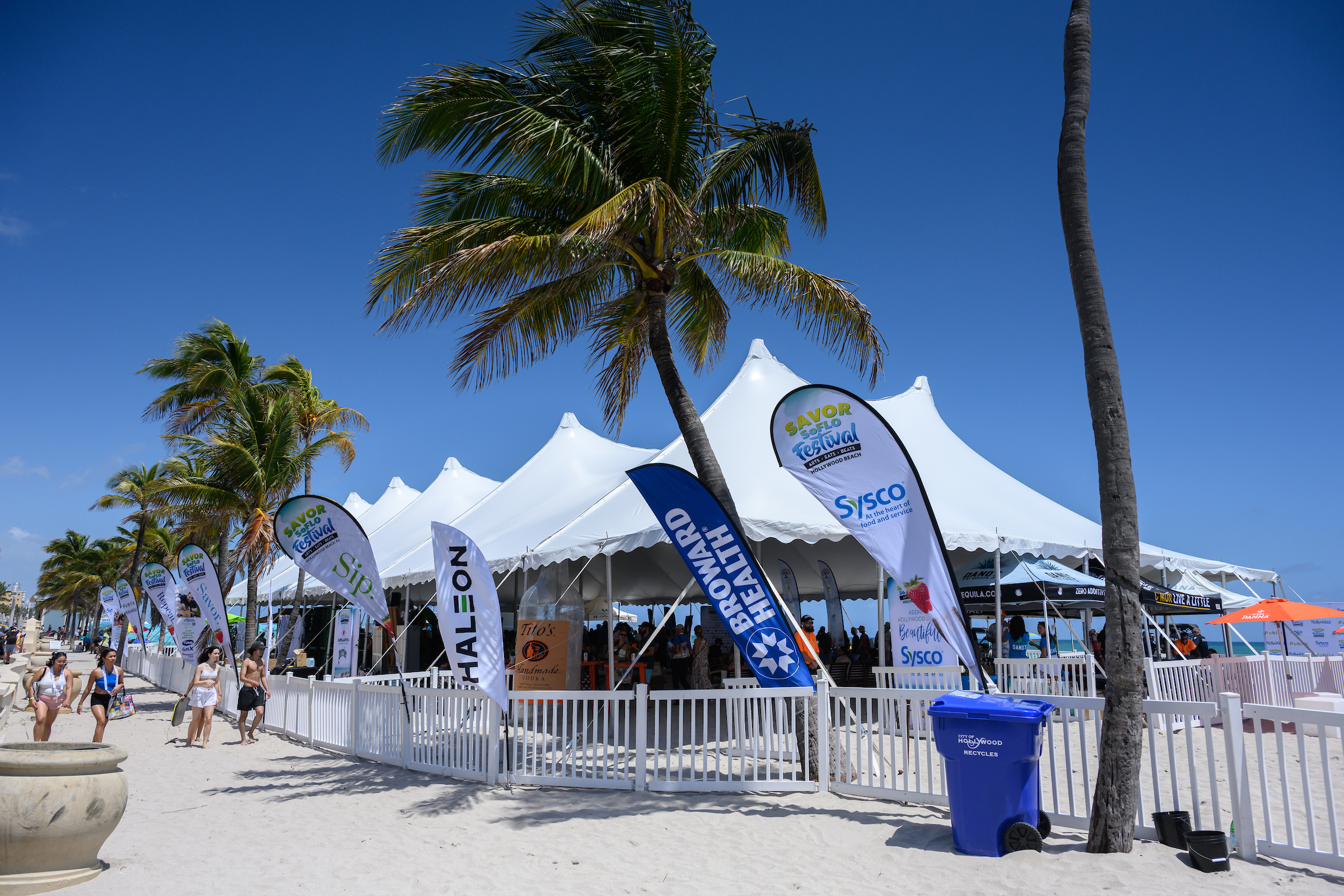 White event tents with sponsor flags line Hollywood Beach under clear blue skies and palm trees, as people stroll by and a blue recycling bin sits in the foreground at SAVOR SoFlo. Lifestyle