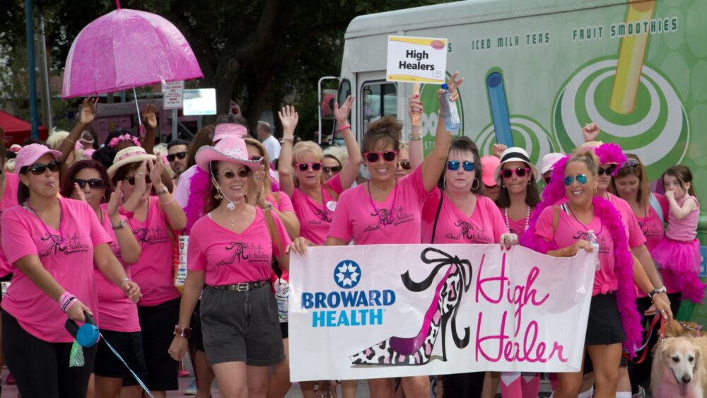 A group of women in matching pink shirts, hats, and sunglasses march together holding a "High Healers" banner for Broward Health at an outdoor event, celebrating Twenty Years in Heels while raising awareness and carrying pink accessories. Lifestyle