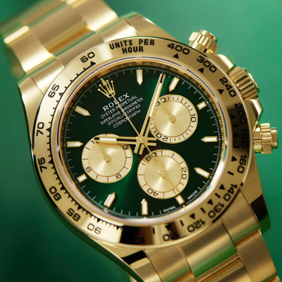 A close-up of a gold Rolex Oyster Perpetual Cosmograph Daytona watch with a green dial, gold hands and hour markers, and three subdials for precise frequency tracking, set against a green background. Lifestyle