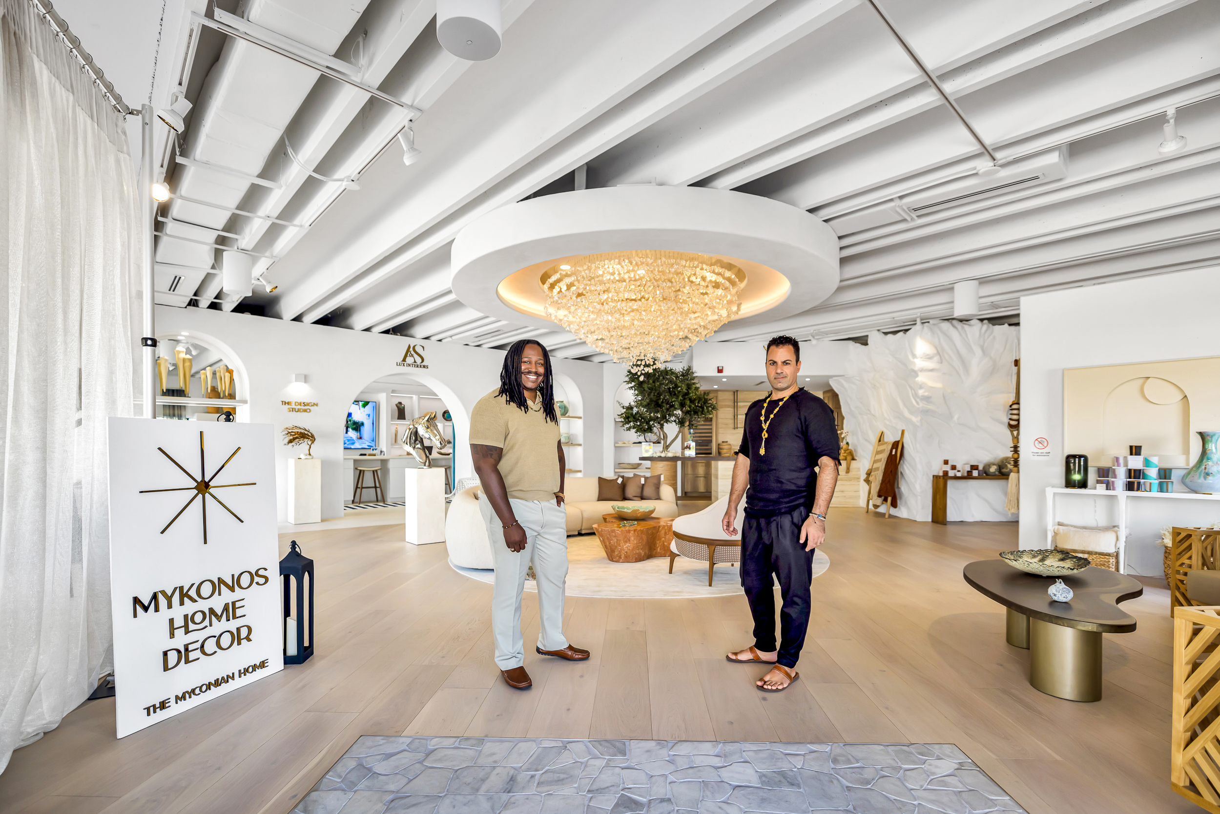 Two men stand smiling in "Mykonos Home Decor," a bright and inviting store featuring modern coastal decor, stylish furniture, unique pieces, and a striking chandelier hanging from the ceiling. Lifestyle
