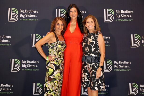 Three women exude Summer Vibes as they smile in front of a Big Brothers Big Sisters of Broward County banner. Two wear patterned dresses, while one makes a BIG Impact in a bright red jumpsuit. Lifestyle