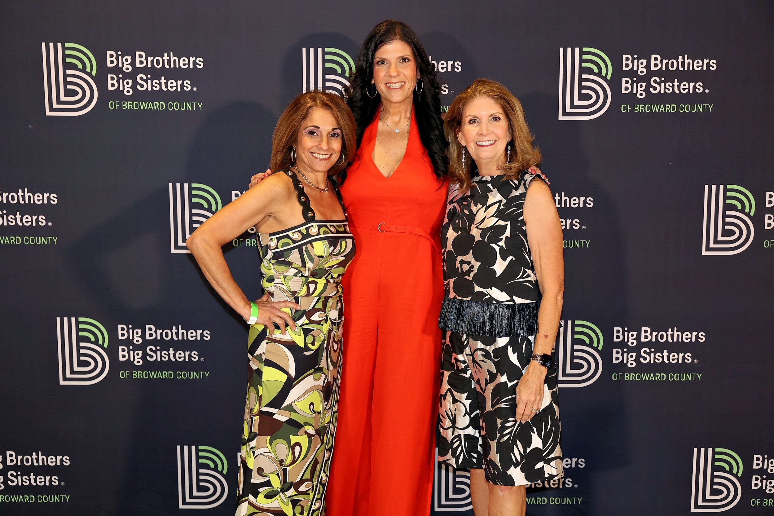 Three women exude Summer Vibes as they smile in front of a Big Brothers Big Sisters of Broward County banner. Two wear patterned dresses, while one makes a BIG Impact in a bright red jumpsuit. Lifestyle