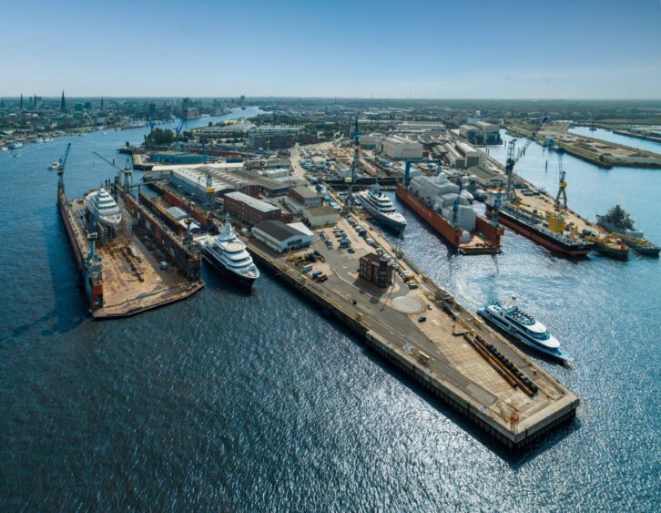 Aerial view of a busy shipyard with several large ships docked for repair and construction—an industrious scene where cranes and warehouses stand as century marks against blue water and a distant city skyline. Lifestyle