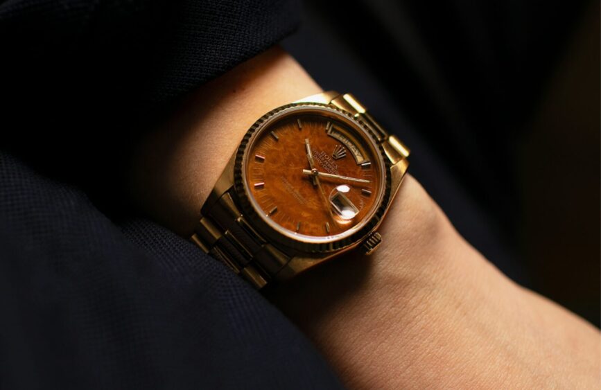 A close-up of a gold wristwatch with a brown wood-textured dial and metal bracelet, showcasing elegant wearable tech on a person's wrist against a dark fabric background. Lifestyle