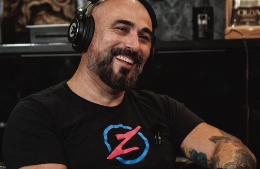 A smiling man with a beard and shaved head wears headphones and a black t-shirt with a colorful "Z" logo, sitting indoors with a microphone clipped to his shirt, ready to discuss topics like Flesh and Canvas. Lifestyle