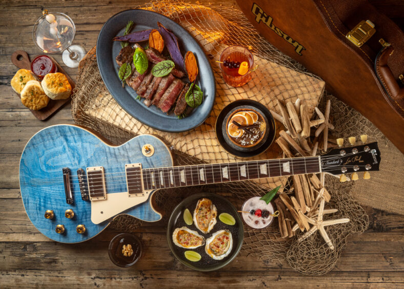 A blue electric guitar sits among plates of steak, oysters with lime, biscuits, and cocktails, atop a rustic wooden surface with decorative netting—blending coastal decor vibes with a Gibson guitar case for a modern coastal touch. Lifestyle