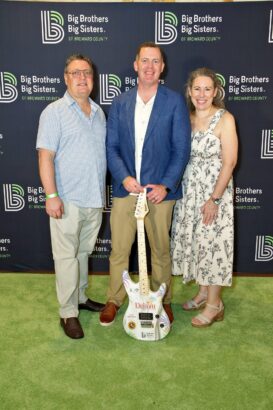 Three adults stand and smile in front of a Big Brothers Big Sisters of Broward County backdrop, radiating summer vibes. The man in the center holds an electric guitar, while the other two stand on either side on a green carpet. Lifestyle