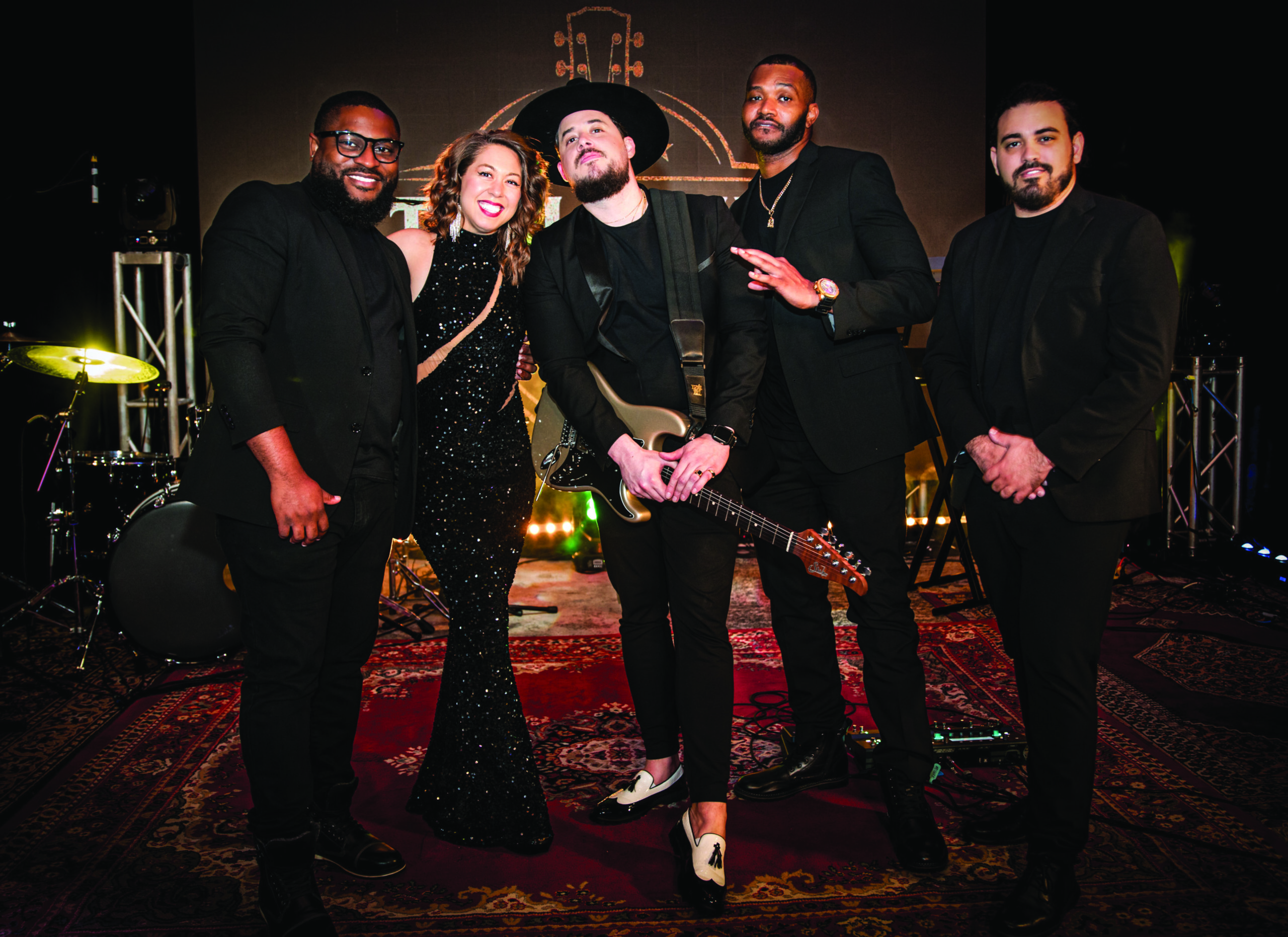 Five people dressed in black, four men and one woman, stand together on a stage in Flagler Village with musical instruments in the background. One man holds an electric guitar; all are smiling and posing to capture the rhythm of the moment. Lifestyle