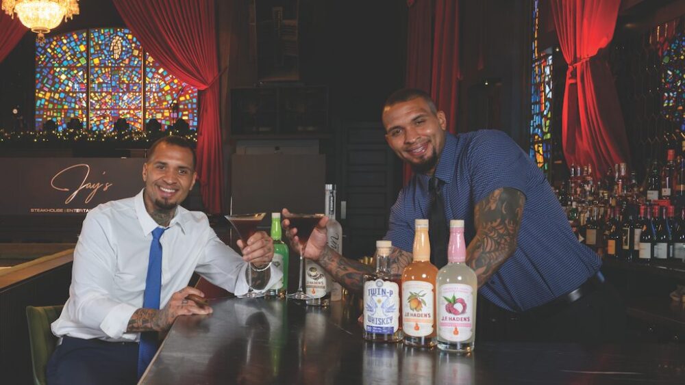 Two men smiling at a bar, holding cocktails. One is seated in a white shirt and blue tie; the other, standing in a blue shirt, pours a drink. Colorful bottles, stained glass windows, and music reminiscent of Hall & Oates set the scene. Lifestyle