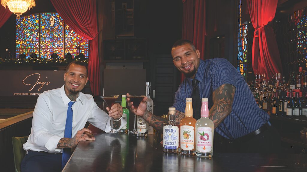 Two men smiling at a bar, holding cocktails. One is seated in a white shirt and blue tie; the other, standing in a blue shirt, pours a drink. Colorful bottles, stained glass windows, and music reminiscent of Hall & Oates set the scene. Lifestyle