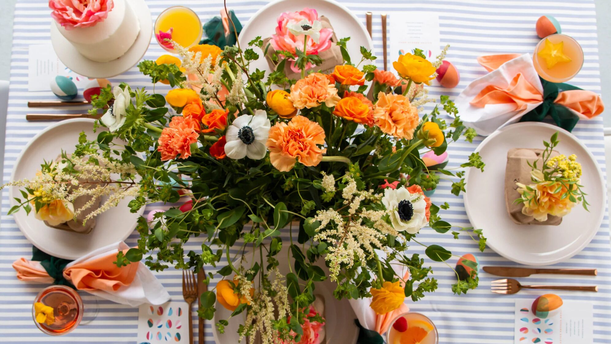 A festive table setting with a striped tablecloth, a colorful floral centerpiece, wrapped gifts, painted eggs, and place settings with napkins and cutlery, creating a vibrant spring or Easter celebration atmosphere. Lifestyle