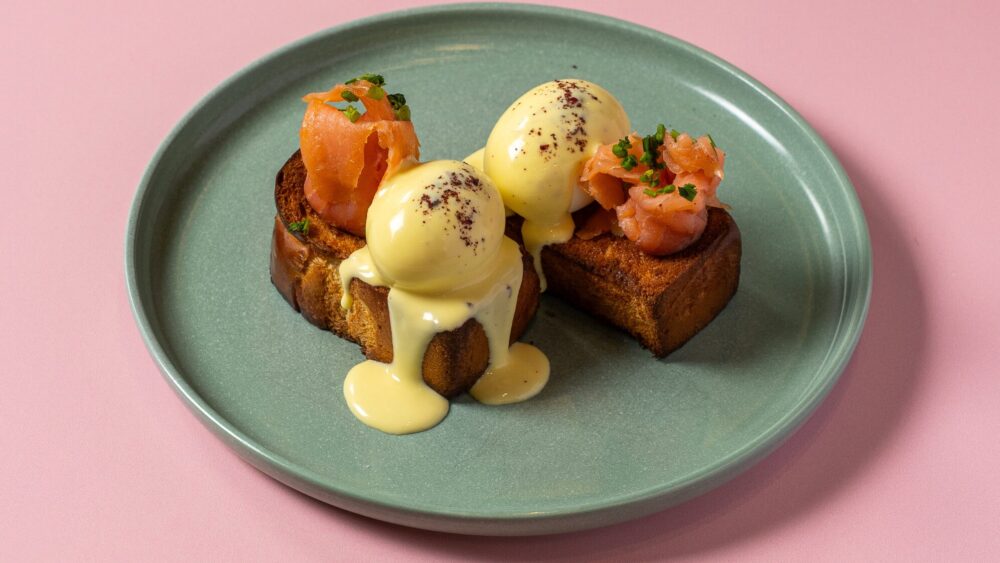 Two pieces of toasted bread topped with poached eggs, hollandaise sauce, and smoked salmon, garnished with herbs—perfect for Easter dining and served on a green plate against a pink background. Lifestyle