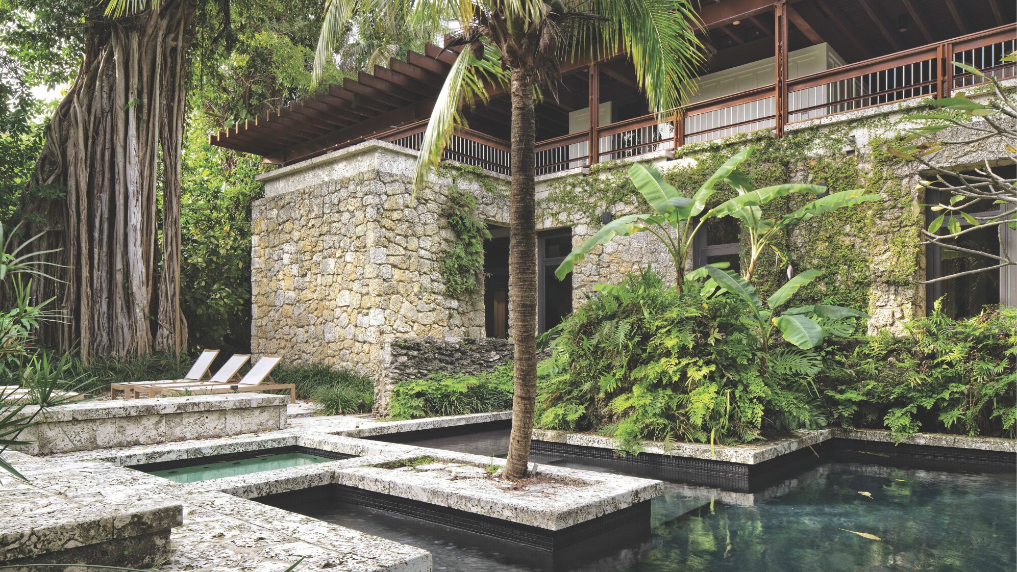 A lush courtyard with a small stone pool, tropical plants, and palm trees surrounds a rustic stone building with a wooden balcony—perfect for savoring small town living. Two lounge chairs sit near the pool, creating a tranquil, inviting atmosphere. Lifestyle