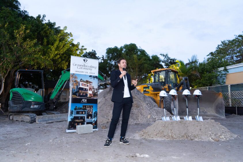 Valentin Carbonell, dressed in a suit, speaks into a microphone at a construction site. Behind him are excavators, hard hats, shovels on a pile of dirt, and a sign announcing the groundbreaking ceremony dated January 20, 2023. Lifestyle