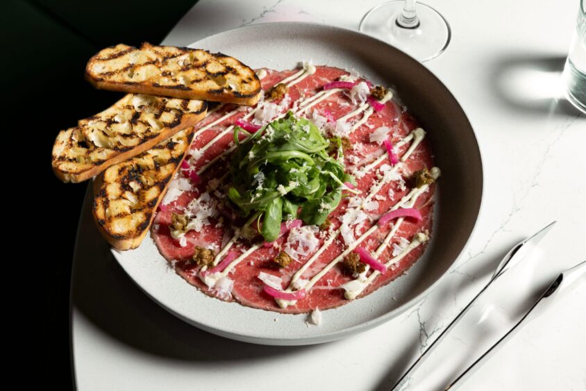 A plate of beef carpaccio topped with arugula, pickled onions, grated cheese, and drizzled sauce—an April Flavor Files highlight—served with three slices of grilled bread on a white table next to a glass of water and a knife. Lifestyle