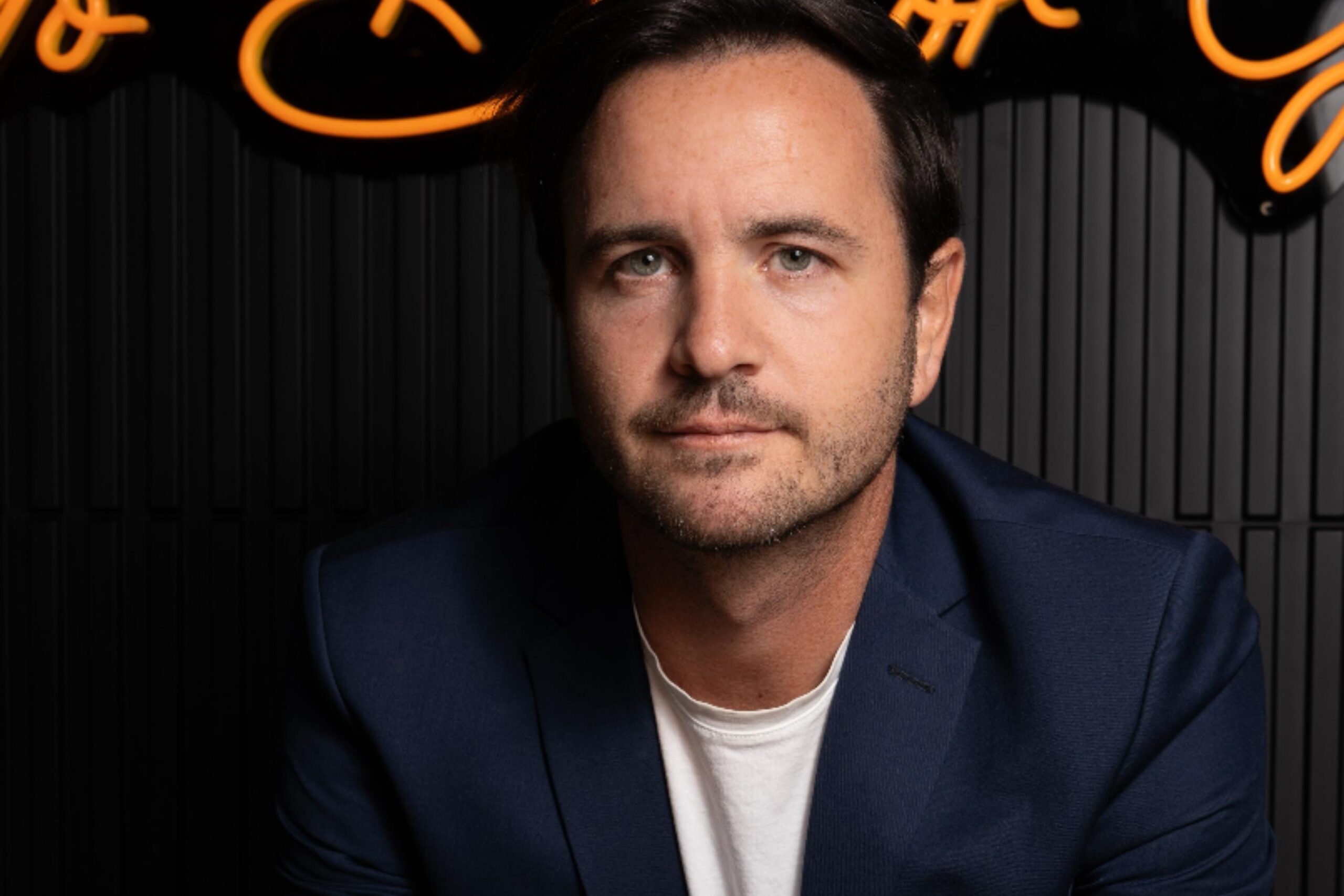 Valentin Carbonell, a man with short dark hair, wearing a navy blazer and white shirt, looks at the camera against a dark, ribbed background illuminated by orange neon lights above him. Lifestyle