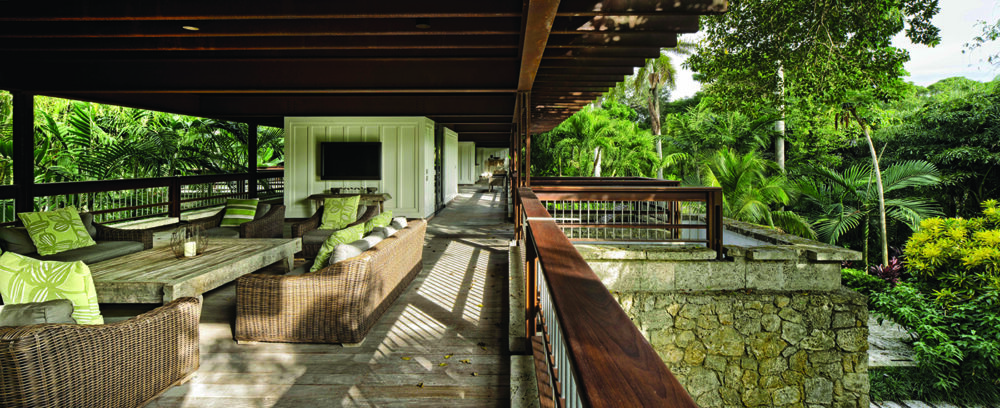 A spacious, covered outdoor patio with wicker furniture and green cushions overlooks lush tropical gardens. Wooden railings and stone columns add rustic Florida charm to the architecture of this inviting setting. Lifestyle