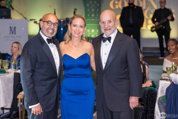 Three people dressed formally in evening wear stand together and smile at a Golden Angels gala event. A woman in a blue strapless dress is between two men in tuxedos, with other guests seated at decorated tables in the background. Lifestyle