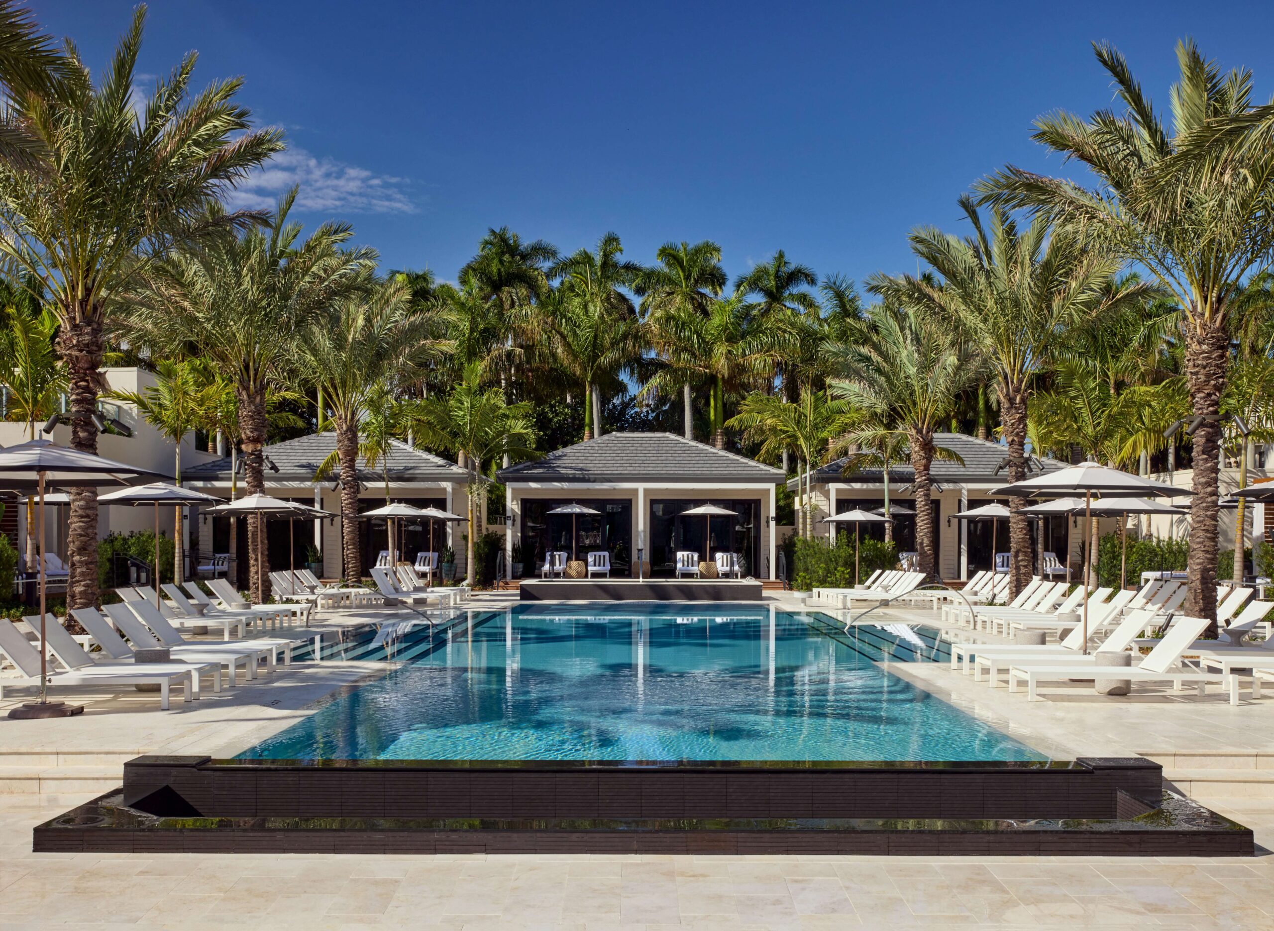A luxurious outdoor pool at the Boca Hotel, surrounded by white lounge chairs and umbrellas, with palm trees and cabanas in the background under a clear blue sky. Lifestyle