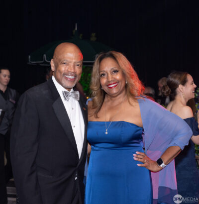 A smiling man in a black tuxedo and bow tie stands next to a smiling woman in a blue off-the-shoulder gown—radiating the elegance of Golden Angels—at a formal event, with other people in formal attire in the background. Lifestyle