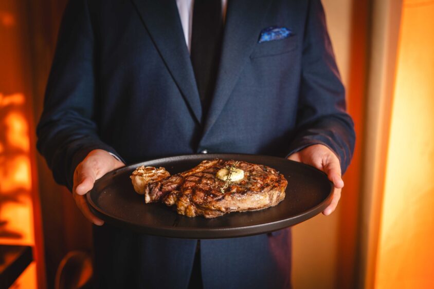 In Wynwood, a person in a dark suit and tie holds a black plate with a grilled steak topped with butter and roasted garlic, as the moon rises over the warmly lit background. Lifestyle