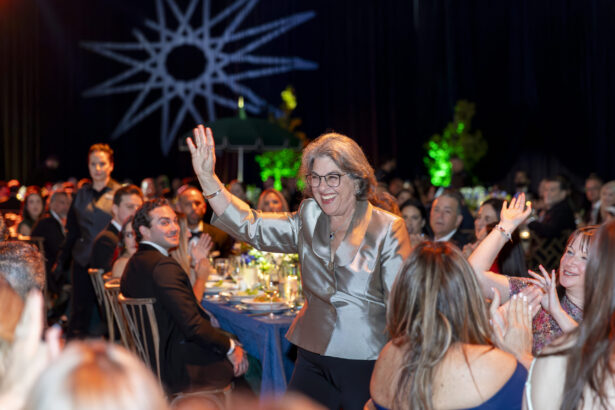 A smiling woman in a silver blazer stands and waves amid a seated crowd at a formal event, with people clapping and colorful lights illuminating the background—a true Golden Angels moment making Real Impact. Lifestyle